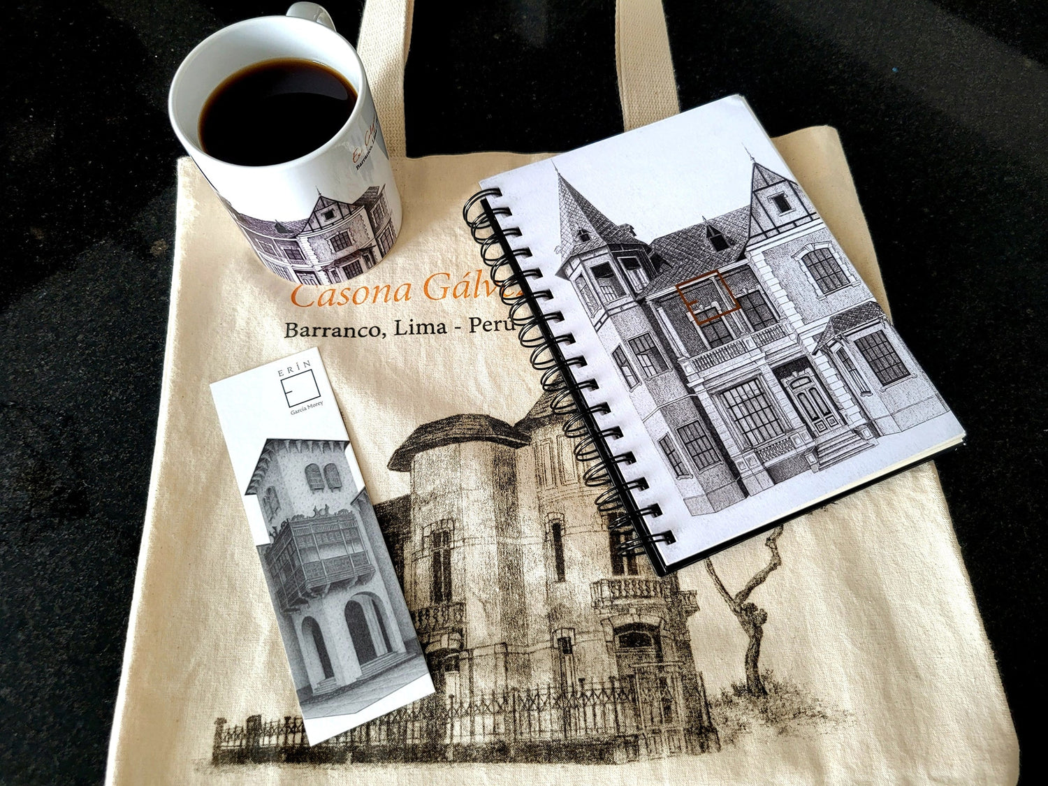 Flat lay con tote bag de tocuyo, libreta de notas con diseño barranquino, taza de café y separador de libro con ilustraciones de Erin García Morey.